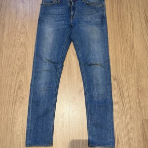 Blå slim jeans från Nudie - Snygga blå slim jeans från Nudie med klassisk femficksdesign och orangea sömmar på bakfickorna. Jeansen har en smal passform och är tillverkade i mjukt denimtyg som sitter tajt längs benen. Perfekta för dig som gillar en stilren och modern look.
