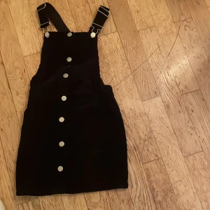 Svart manchesterklänning från Lindex - Supersnygg svart manchesterklänning från Lindex med justerbara axelband och silvriga knappar framtill. Klänningen har en cool, rak passform och är perfekt att styla med en t-shirt under. Materialet är mjukt och ger en chill vibe.