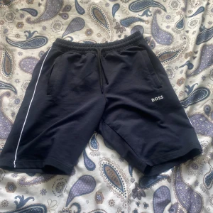 Svarta shorts från Hugo Boss - Svarta shorts från Hugo Boss i 100% bomull med vit rand längs ena benet och broderad BOSS-logga framtill. De har elastisk midja med snörning och två sidofickor, samt en bakficka. Perfekta för chill dagar eller träning.