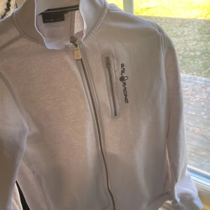 Vit vindjacka från Sail Racing - Fräsch vit vindjacka från Sail Racing med stilren design och logga på bröstet. Jackan har en praktisk bröstficka med dragkedja, hel dragkedja framtill och hög krage. Perfekt för dig som gillar sportig och clean look.