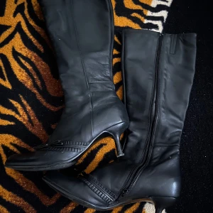 Svarta stövlar med klack och dragkedja - Säljer ett par svarta stövlar i skinn med smal klack och dragkedja på insidan. Snygga detaljer framtill med diskret rosett och ribbad kant. Perfekta för dig som vill ha en klassisk och stilren look med lite edge.