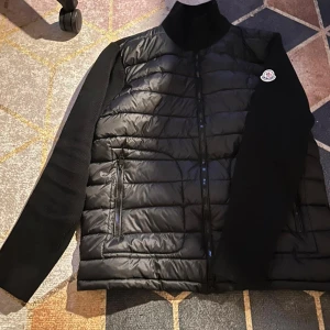 Svart pufferjacka från Moncler - Säljer en svart pufferjacka från Moncler med stickade ärmar och hög krage. Jackan har quiltad framsida, dragkedja och två sidofickor med zip. Moncler-logga på vänster ärm och coolt serietryck på insidan. Perfekt för dig som gillar stilren och sportig look.