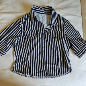 Randig skjorta med trekvartsärm - Snygg skjorta med svart och vit randigt mönster, klassisk krage och två bröstfickor. Skjortan har trekvartsärm och knappar framtill. Perfekt för en stilren och trendig look.