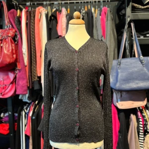 Glittrig svart kofta - Snygg svart ribbad kofta från Joy med glittriga inslag och lila knappar. Koftan har v-ringning och långa ärmar. Materialet är stretchigt och formar sig efter kroppen. Storleken är liten så skulle säga att den mer är en M