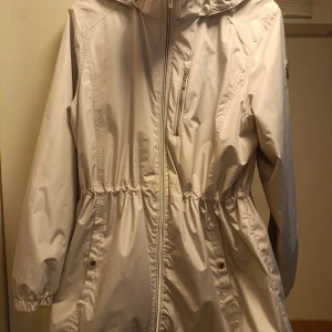 Ljusgrå vindjacka från Tuxer, stl L - Snygg ljusgrå vindjacka från Tuxer med huva, dragkedja framtill och praktiska fickor. Jackan har resår i midjan och vid ärmsluten för en skön passform. Perfekt för blåsiga dagar och enkel att matcha med din stil.