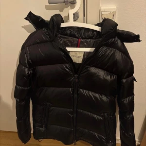 Svart Moncler dunjacka med huva - Snygg svart dunjacka från Moncler med glansig finish och huva. Jackan har dragkedja framtill, två fickor med dragkedja och klassisk Moncler-logga på ärmen. Perfekt för kalla dagar och ger en riktigt clean look. Är i storlek 4 byten är intressant 