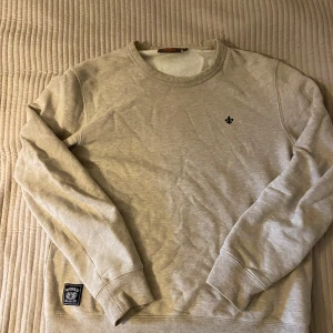 Beige sweatshirt från Morris Stockholm - Säljer en stilren beige sweatshirt från Morris Stockholm med rund halsringning och diskret logga på bröstet. Tröjan har ribbade muddar och en patch nertill. Perfekt för dig som gillar klassisk och enkel stil.