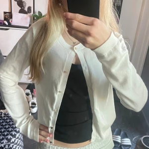 Vit kofta från Lindex med knappar - Säljer en klassisk vit kofta från Lindex i storlek 158/164. Koftan har rund halsringning, långa ärmar och stängs med små knappar framtill. Perfekt att slänga över en topp eller klänning för en clean och enkel look. Använt 1 gång 