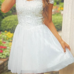 Vit kort klänning med spets och pärlor - Superfin vit kort klänning med spets och broderade blommor upptill, dekorerad med små pärlor och paljetter. Kjolen är i flera lager tunn tyll som ger en fluffig känsla. Klänningen har snörning i ryggen för perfekt passform och en romantisk vibe.