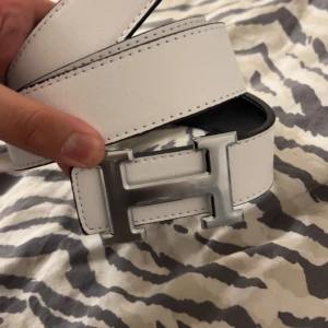 Vitt och svart skärp med H-spänne - Säljer ett snyggt bälte i vitt och svart skinn med ett stort, silvrigt H-spänne. Bältet är vändbart och har stilrena sömmar längs kanterna. Perfekt accessoar för att lyfta din outfit med en lyxig touch.