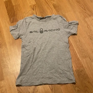 Grå Sail Racing t-shirt i bomull - Snygg grå t-shirt från Sail Racing med svart logga tryckt på bröstet. Klassisk rund hals och korta ärmar. Tillverkad i mjuk bomull som är skön att ha på sig. Perfekt för en avslappnad och sportig stil.