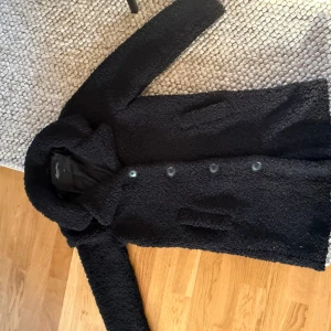 Kappa  - Mysig svart teddyjacka från Happy Holly med klassisk krage och knäppning framtill. Jackan har två fickor och är tillverkad i ett fluffigt teddy-material som håller dig varm under kyliga dagar. Perfekt för dig som gillar en stilren och trendig look.