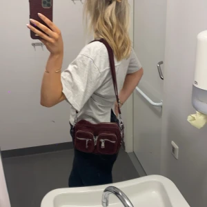 Vinröd axelväska med dragkedjor - Säljer en vinröd axelväska i skinn med två framfickor och silvriga dragkedjor. Väskan har justerbar axelrem och en kompakt, fyrkantig form som rymmer det viktigaste. Perfekt för dig som vill ha en snygg och praktisk väska till vardagen.