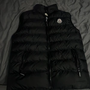 Svart dunväst från Moncler, S priset kan diskuteras - Svart dunväst från Moncler i storlek S. Västen har quiltad design, hög krage och två dragkedjeförsedda fickor. Moncler-logga på bröstet och coolt serietidningsmärke på insidan. Perfekt för lager-på-lager och streetstyle.