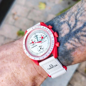 Ny i box Omega x Swatch MoonSwatch röd/vit keramisk klocka - Helt ny Omega x Swatch MoonSwatch Speedmaster - Mission to Mars i låda inkl. allt. Endast använd 1 gång för foto. Klockan är keramisk och har vit urtavla med röda och gröna visare, tachymeter-skala och flera subdials. Armbandet är vitt med Omega-logga och texten Speedmaster. 