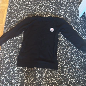 Svart långärmad tröja från Moncler - Svart långärmad tröja från Moncler med klassisk rund hals och broderad logga på bröstet. Tröjan har en stilren och enkel design som passar perfekt till jeans eller dressade byxor. Mjuk och skön känsla, perfekt för kyligare dagar.