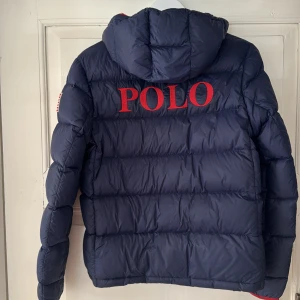 Mörkblå dunjacka Polo Ralph Lauren XL - Mörkblå dunjacka från Polo Ralph Lauren med röd dragkedja och detaljer. Jackan har huva, två fickor med dragkedja och stor röd POLO-logga broderad på ryggen. Klassisk pufferstil, perfekt för kalla dagar.