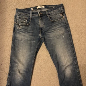 Replay Anbass jeans Aged 05 33/34 med slitningar - Snygga blå Replay jeans i storlek 33/34 men sitter mer som 32/33 med unika slitningar och tvättade detaljer. Klassisk femficksmodell med ljusare partier och slitna kanter på fickorna. Tillverkade i bomull med hållbarhetsfokus. Perfekt för dig som gillar en avslappnad och trendig look.