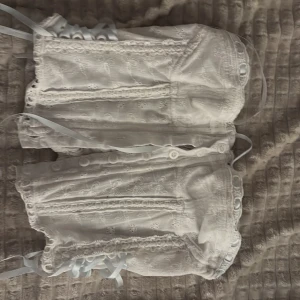 Vit spetskorsett med snörning - Superfin vit korsett med broderade spetsdetaljer och ljusblå snörning längs sidorna. Modellen är croppad och har dekorativa knappar framtill. Materialet är bomull med spets och broderade blommor för en romantisk vibe. Perfekt för dig som vill ha en unik och söt topp.