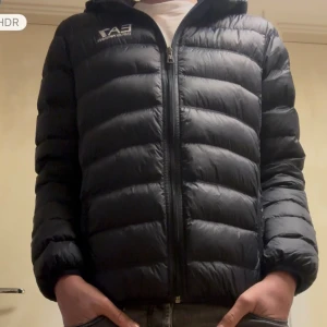 Svart pufferjacka EA7 Emporio Armani - Snygg svart pufferjacka från EA7 Emporio Armani med huva och vit logga på bröstet. Jackan har quiltad design, dragkedja framtill och två sidofickor. Perfekt för kalla dagar och har en modern, sportig look.