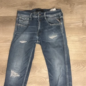Replay Anbass blå jeans, storlek 32 - Snygga Replay Anbass jeans i klassisk blå tvätt med slitningar och distressed detaljer på benen. Modellen har fem fickor, normal passform och raka ben. Jeansen är tillverkade i stretchigt bomullsmaterial för extra komfort och har Replay-logga på bakfickan.