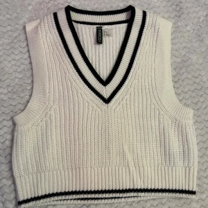 Vit stickad väst från H&M Divided - Snygg vit stickad väst från H&M Divided i storlek XS. Västen har en v-ringad hals med svarta ränder och ribbad kant nedtill. Perfekt att styla över en skjorta eller t-shirt för en trendig look.
