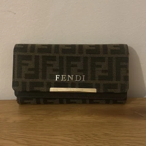 Brun Fendi plånbok - Snygg brun plånbok från Fendi med klassiskt FF-logomönster i mörka toner. Plånboken har flera fack för kort och sedlar, magnetlås och detaljer i guld. Materialet är skinn och textil, och Fendi-loggan syns både på utsidan och insidan.