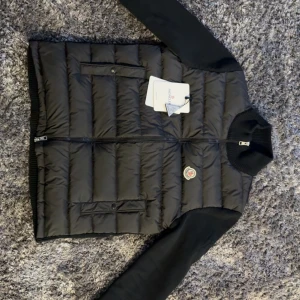 Svart pufferjacka från Moncler - Säljer en svart pufferjacka från Moncler med dunfylld framsida och stickade ärmar och rygg. Jackan har dragkedja framtill, ribbad krage och Moncler-logga på bröstet. Perfekt för dig som vill ha en stilren och varm jacka med snygga detaljer.