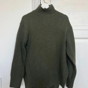 Olivgrön polotröja i ull - Mysig olivgrön polotröja med hög krage och ribbade muddar. Tröjan är stickad i ull och har en loose passform med långa ärmar. Perfekt för kyliga dagar och enkel att styla till jeans eller kjol.