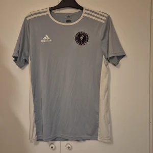 Fort Lauderdale fotbollströja Adidas M - Snygg ljusblå fotbollströja från Adidas med Fort Lauderdale Club de Fútbol-logga på bröstet. Tröjan har vita detaljer på axlar och sidor, samt klassiska Adidas-ränder. Tillverkad i lätt och ventilerande Climalite-material, perfekt för träning eller match.