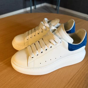 Alexander McQueen vita sneakers med blå detalj - Säljer ett par chunky vita sneakers från Alexander McQueen med blå mockadetalj på hälen och breda vita skosnören. Skorna har en hög plattformsula och diskret logga på både häl och plös. Tillverkade i skinn och mocka för en lyxig känsla.