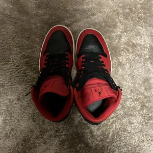 Nike Air Jordan 1 High röd/svart - Nike Air Jordan 1 High i klassisk röd och svart färg med vit sula. Skorna har snörning, rund tå och är tillverkade i skinn med syntetdetaljer. Ikonisk Jordan-logga på sidan och Nike-märke på sulan. Perfekt för dig som vill sticka ut med en legendarisk sneaker.