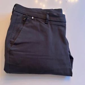 Grå kostymbyxor Boss Kaito Slim Fit i storlek 36 - Snygga grå kostymbyxor från Boss, modell Kaito Slim Fit. Byxorna har klassisk rak passform med smal siluett, bälteshällor och sidofickor med dragkedja. Materialet är mjukt och känns lyxigt, perfekt för dig som gillar stilrena och moderna plagg.