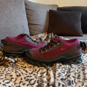 Vinröda chunky sneakers från Nike - Säljer ett par feta vinröda och mörkbruna sneakers från Nike med grov sula och coola detaljer. Skorna har snörning med metallöglor och är gjorda i en mix av textil och syntetmaterial. Perfekta för dig som gillar en unik och robust stil.