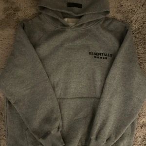 Grå Essentials Fear of God hoodie - Säljer en grå hoodie från Essentials Fear of God med svart logga på bröstet och ryggen. Klassisk oversized passform, stor huva och magficka. Perfekt för dig som gillar streetwear och vill ha en stilren look.