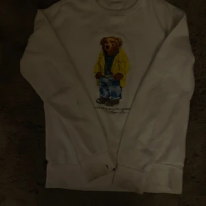 Vit sweatshirt Polo Ralph Lauren björn - Vit sweatshirt från Polo Ralph Lauren med ikonisk björn på bröstet. Tröjan har rund halsringning och långa ärmar. Björnen har gul jacka och jeans, vilket ger en cool och lekfull vibe. Perfekt för dig som gillar streetstyle och klassiska märken.
