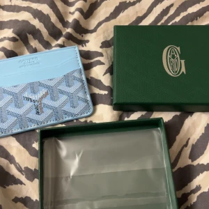 Blå plånbok med mönster - Snygg ljusblå plånbok från med ikoniskt grafiskt mönster och flera fack för kort. Kommer med original grön ask. Perfekt för dig som vill ha något unikt och stilrent i fickan.