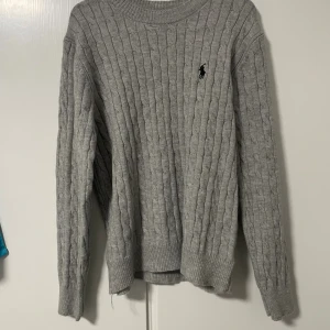  Polo Ralph Lauren tröja  - Nyskick kabelstickad grå tröja från Polo Ralph Lauren med klassisk rund halsringning och broderad logga på bröstet. Tröjan har långa ärmar och ribbade muddar för en snygg passform. Perfekt för lager-på-lager under kyliga dagar.