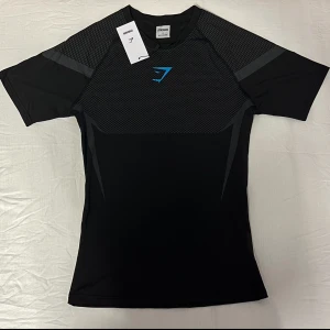 Gymshark Onyx - Svart t-shirt från Gymshark med korta ärmar och sportig passform. Framsidan har ett diskret mönster och en blå Gymshark-logga på bröstet. Materialet är lätt och stretchigt, perfekt för träning. Ett till ett. Snygga grå detaljer på axlar och rygg för extra stil. Denna tröja är limited edition, alltså går det inte att få tag på. Vid någora funderingar är det att bara att fråga 🤗. 