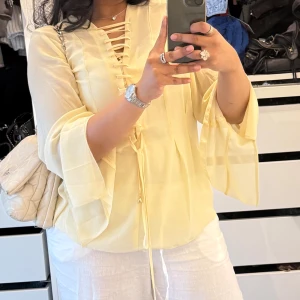 Ljusgul blus med snörning - Säljer en ljusgul blus med v-ringning och snygg snörning framtill. Blusen har vida, fladdriga ärmar och är gjord i ett tunt, luftigt material som känns skönt mot huden. Perfekt för dig som gillar en feminin och trendig look.