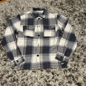 Rutig blå skjorta från KappAhl - Riktigt snygg rutig skjorta från KappAhl i blått, grått och vitt. Skjortan har två bröstfickor med lock, klassisk krage och knappar framtill. Långärmad och tillverkad i mjukt bomullsmaterial, perfekt för en chill och stilren look.