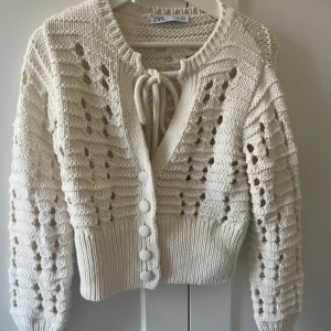 Beige stickad kofta från Zara - Superfin beige stickad kofta från Zara med snyggt hålmönster, knytning vid halsen och stora knappar framtill. Modellen är croppad med långa ärmar och breda muddar. Perfekt att slänga över en topp för en chill look.