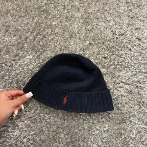 Mörkblå mössa från Polo Ralph Lauren - Snygg mörkblå mössa från Polo Ralph Lauren med klassisk röd broderad logga framtill. Mössan är ribbstickad i nederkant och tillverkad i mjukt material som håller dig varm under kalla dagar. Perfekt accessoar för en stilren look.