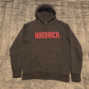 Svart hoodie från Hoodrich med röd logga - Svart hoodie från Hoodrich i storlek medium med stor röd HOODRICH-logga på bröstet. Klassisk känguruficka framtill, huva med snörning och ribbade muddar.