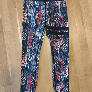 Fjädermönstrade leggings från Stronger - Snygga leggings från Stronger med färgstarkt fjädermönster i svart, rött, blått och vitt. Hög midja och tight passform, perfekta för träning. Tillverkade i stretchigt syntetmaterial som andas och sitter skönt under hela passet.
