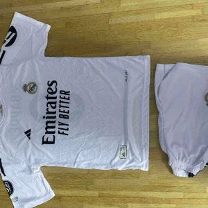Real Madrid Mbappé fotbollströja vit - Säljer en vit Real Madrid fotbollströja från Adidas med Mbappé och nummer 9 på ryggen. Tröjan har korta ärmar, klubbmärke på bröstet och Emirates Fly Better-tryck framtill. Tillverkad i lätt och ventilerande material, perfekt för match eller träning.