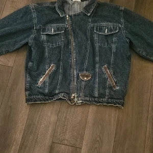 Oversized blå jeansjacka med dragkedja - Säljer en oversized blå jeansjacka med dragkedja framtill, två bröstfickor med lock och snedställda fickor med läderdetaljer. Jackan har klassisk krage och långa ärmar. Perfekt för dig som gillar en avslappnad och retro stil.