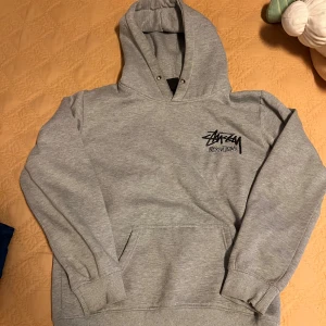 Grå Stüssy hoodie med logga - Säljer en grå hoodie från Stüssy med stor svart logga på ryggen och mindre logga på bröstet. Klassisk känguruficka, ribbade muddar och huva. Perfekt för dig som gillar streetwear och vill ha en clean look.
