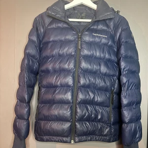 Blå pufferjacka från Peak Performance 160 - Marinblå pufferjacka från Peak Performance med huva och dragkedja framtill. Jackan har två dragkedjeförsedda fickor och ribbade muddar vid ärmsluten. Perfekt för kyliga dagar och har en klassisk pufferstil. Jackan har ett litet hål men täcks ganska bra av luvan. 