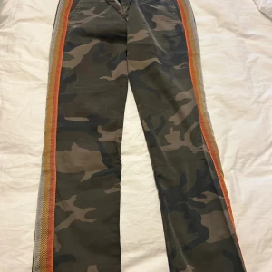 Camo cargopants från Please med revärer - Snygga cargopants från Please i camouflage-mönster med coola orange och gula revärer längs sidorna. Byxorna har raka ben, normal passform och är tillverkade i bomull med lite stretch. Bakfickor med dekorativt hjärtdetalj. Perfekta för en streetstyle-look.